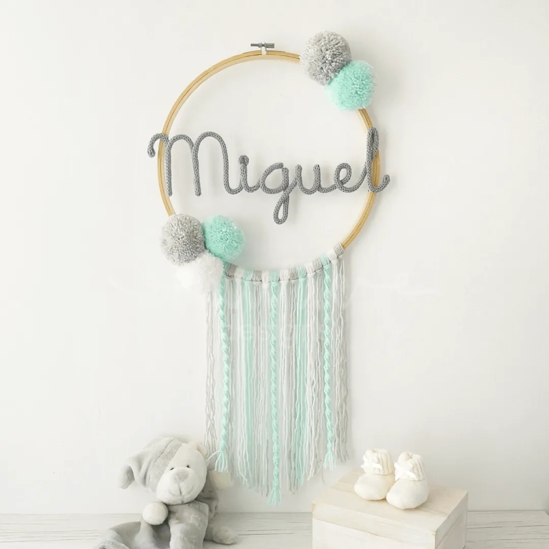 Caça sonhos artesanal em bastidor de madeira natural com nome personalizado, decorado com pompons e fios de lã, ideal para a decoração de quarto infantil