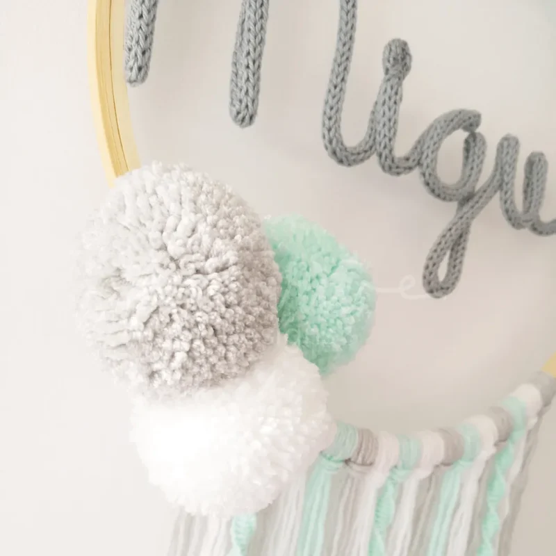 Caça sonhos artesanal em bastidor de madeira natural com nome personalizado, decorado com pompons e fios de lã, ideal para a decoração de quarto infantil