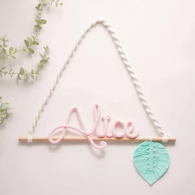 Decoração infantil com o nome personalizado em crochet, pendurado num suporte de madeira com corda em macramé e uma folha decorativa verde em macramé