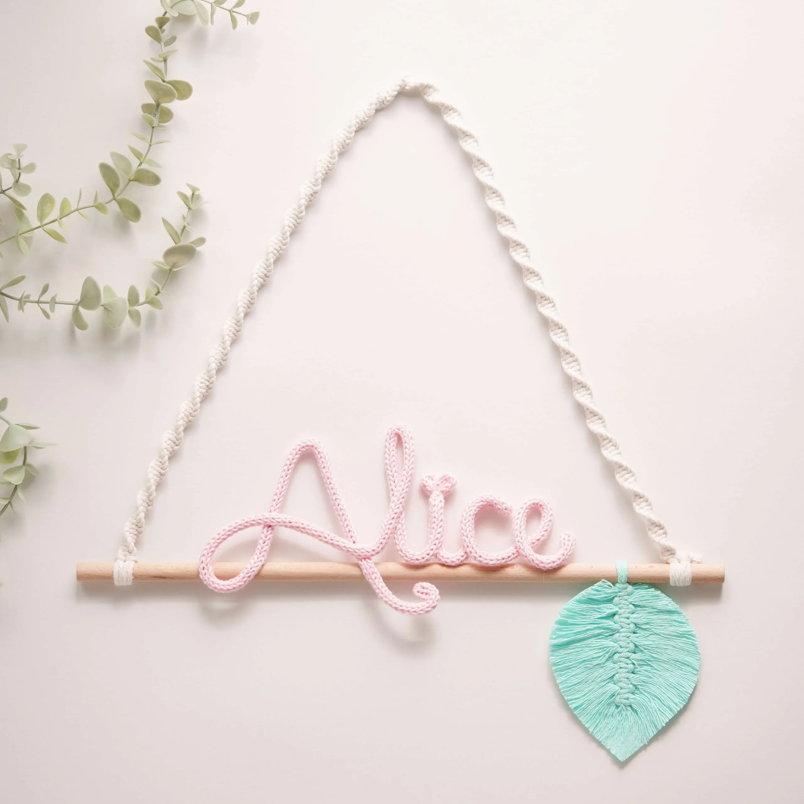Decoração infantil com o nome personalizado em crochet, pendurado num suporte de madeira com corda em macramé e uma folha decorativa verde em macramé