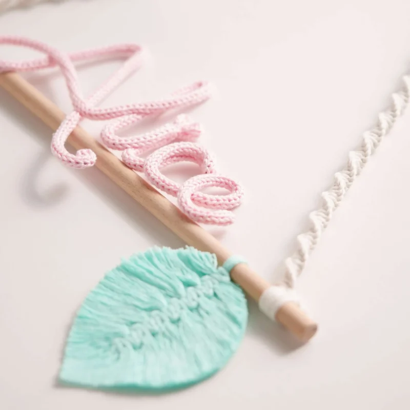 Decoração infantil com o nome personalizado em crochet, pendurado num suporte de madeira com corda em macramé e uma folha decorativa verde em macramé